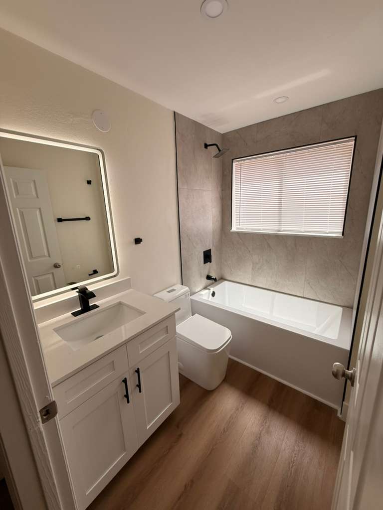 Guest bathroom remodel with a shaker vanity, backlit mirror, matte black fixtures and a tiled tub surround